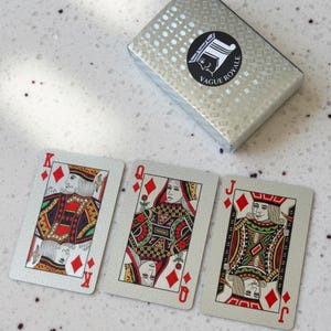 Puede incluir: Una baraja de cartas con una caja plateada. Se ven tres cartas: el Rey, la Reina y la Jota de diamantes. Las cartas tienen un fondo plateado con detalles rojos, negros y dorados. La caja tiene el texto "Vague Royale".