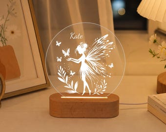 May include: A circular acrylic night light featuring a white etched design of a fairy with wings and butterflies, personalized with the name Kate. The clear acrylic disc is mounted on an oval wooden base that illuminates the etched graphic.