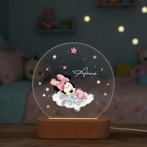 Puede incluir: Luz de noche redonda con base de madera, con Minnie Mouse durmiendo en una nube con un conejito. El nombre "Anna" está escrito en la parte delantera. El diseño incluye estrellas y detalles rosas.