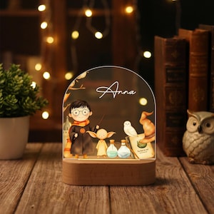 May include: A decorative night light featuring a cartoon illustration of a boy with glasses, a scarf, and a wand, along with other characters. The name "Anna" is written on the clear acrylic panel. The light sits on a wooden base.