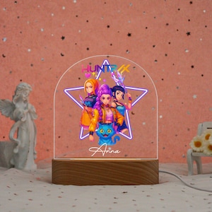 Op de afbeelding: Een gepersonaliseerde LED-nachtlamp met een cartoonillustratie van drie vrouwelijke personages en een blauwe kat. Het ontwerp is geplaatst in een stervormig frame, met de naam "Anna" eronder. Het licht staat op een houten voet.