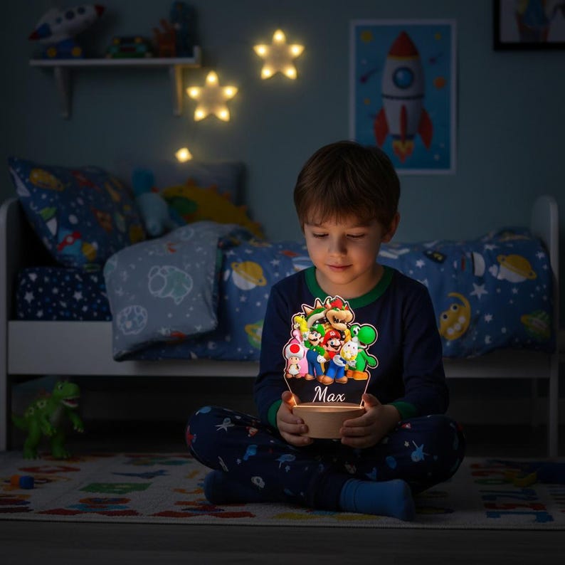 May include: A child holds a glowing night light featuring a colorful Super Mario Bros. design. The light has the name "Max" on it. The child is wearing blue pajamas and is sitting on a patterned rug in a bedroom with star-shaped lights.