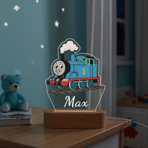 Könnte beinhalten: Blau-rotes Nachtlicht in Form einer Lokomotive mit dem Namen "Max" auf einem Holzsockel. Die Lokomotive hat ein lächelndes Gesicht und die Nummer "1". Der Hintergrund ist eine hellblaue Wand mit Sternaufklebern.