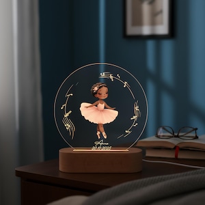May include: A personalized night light featuring a ballerina in a pink tutu, surrounded by musical notes. The design is etched on a clear acrylic circle, illuminated by a warm light from a wooden base. The name "Anna" and a date are also included.