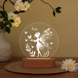 May include: A personalized fairy night light with the name "Kate" etched on a clear, circular acrylic panel. The design features a fairy silhouette with butterflies and foliage, set on a wooden base. The light emits a soft glow.