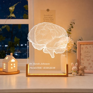 May include: A clear acrylic lamp featuring a white wireframe brain illustration, mounted on a rectangular wooden base. The base displays the text Dr. Sarah Johnson and PEDIATRIC SURGEON in white lettering. The lamp emits a warm light.