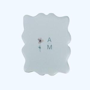 May include: A light blue, wavy-edged coaster with a floral design and the letters "A M" in a gray font. The coaster has a smooth, glossy finish and a minimalist aesthetic.