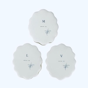 May include: Three off-white, scalloped-edge coasters. Each coaster has a letter (M, L, or V), the text "atelier_03", and a small floral illustration in a muted blue-gray color. The coasters are arranged on a white background.