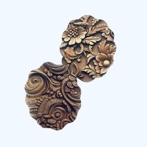 May include: Two intricately carved wooden plaques. One features floral designs, including large flowers and leaves. The other has a swirling, ornate pattern. Both are oval-shaped and have a warm, brown color.