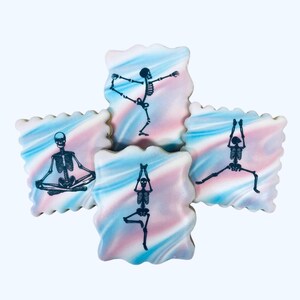 May include: Square-shaped cookies with a scalloped edge, decorated with a blue, pink, and white swirled pattern. Each cookie features a black skeleton in a different yoga pose. The cookies are arranged in a cross shape.