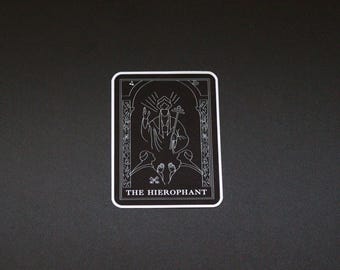 May include: A black and white tarot card sticker featuring "The Hierophant." The card design includes an illustration of a robed figure with a staff, flanked by two figures. The card has a white border.