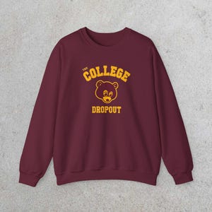Puede incluir: Sudadera granate con el texto "THE COLLEGE DROPOUT" en amarillo, junto con un gráfico de oso de dibujos animados. La sudadera tiene mangas largas y cuello redondo. El diseño está centrado en la parte delantera.