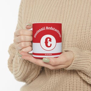 May include: White ceramic mug with a red and white design. The mug features the text "Cincinnati Redstockings" and a large white letter "C" in a red circle. The text "1890" is printed below the circle.