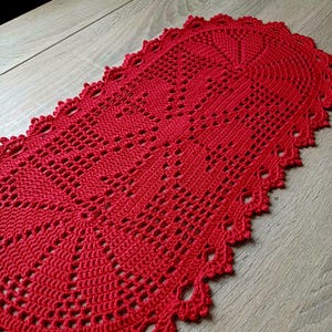 Puede incluir: Un camino de mesa rojo vibrante, tejido a mano con ganchillo. El camino de mesa presenta un intrincado diseño calado con un borde festoneado. El textil hecho a mano se muestra sobre una superficie de madera clara, mostrando la artesanía detallada.
