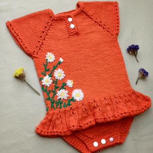 Puede incluir: Mono de bebé de punto naranja con un diseño de bordado floral de margaritas blancas. El mono tiene un dobladillo con volantes y cierres a presión.