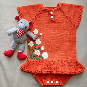 Puede incluir: Mono de bebé de punto naranja con bordado de margaritas blancas y un dobladillo con volantes. El mono tiene un cierre de botón en el cuello y broches en la parte inferior.