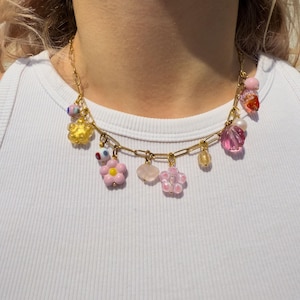 Può includere: Una collana a catena dorata ornata di ciondoli colorati. I ciondoli includono forme di fiori rosa, un fiore giallo, un cuore rosa e varie perline rosa, gialle e trasparenti. La collana è indossata su uno sfondo bianco.