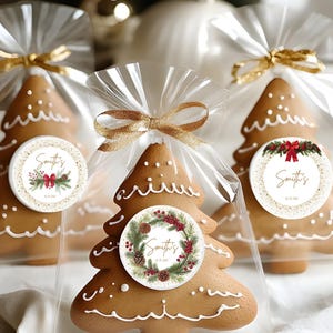 Puede incluir: Galletas de jengibre con forma de árbol de Navidad en bolsas de celofán transparente atadas con cinta dorada. Cada galleta tiene detalles de glaseado blanco y una pegatina redonda con texto y un diseño de corona festiva. Las galletas están sobre una superficie blanca.