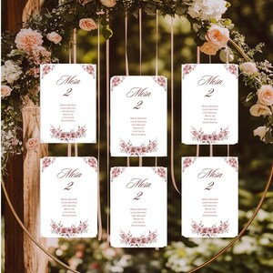 May include: Six white table assignment cards with floral accents and the word "Mesa 2" are suspended from a gold hoop adorned with flowers. Each card lists names below the table number, ideal for wedding or event decor.