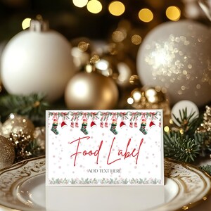 Puede incluir: Una tarjeta blanca para etiquetas de alimentos con la inscripción roja "Food Label" y "Add Text Here". La tarjeta está decorada con calcetines navideños, gorros de Papá Noel y copos de nieve. La tarjeta está sobre un plato blanco con borde dorado, con adornos navideños en el fondo.