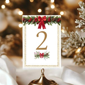 以下が含まれることがあります： 金色のテーブルナンバーホルダー。白いカードには数字の「2」が描かれています。カードは赤いリボン、松ぼっくり、緑で飾られています。ホルダーは丸いベースと上部にループが付いています。