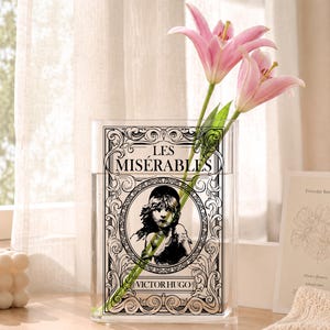 May include: A clear rectangular glass vase featuring a black and white graphic print of the book cover for Les Misérables by Victor Hugo, including an illustration of a young person, with two pink lily flowers placed inside the vase.