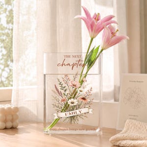 Pode incluir: Um vaso retangular de vidro transparente com uma ilustração impressa de um buquê de flores, o texto "THE NEXT chapter", o nome "EMILY" em uma faixa e a frase "every chapter brings new growth" na base, contendo dois lírios cor-de-rosa.