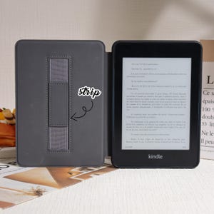 May include: A black Kindle e-reader with a gray cover, open to display text. The cover has a gray elastic strip for securing the device. The Kindle is on a light surface, with a paper bag visible in the background.