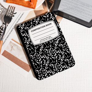 May include: A black and white composition book with a speckled pattern. The cover features a white label with the words "COMPOSITION BOOK". The book measures 24.7 x 19.0 cm and contains 100 sheets. A Kindle e-reader is visible in the background.