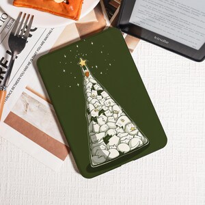 May include: A green phone case featuring a Christmas tree design made of white and gray cats. A gold star tops the tree, with small stars scattered around. The case is on a white surface with a fork and a Kindle.