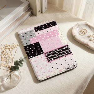 Può includere: Una custodia per tablet a fantasia con un design patchwork con quadrati rosa, neri, grigi e bianchi. I quadrati sono decorati con stelle, fiocchi e altri motivi. La custodia è su una superficie chiara.