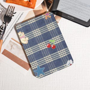 May include: Blue plaid Kindle cover with embroidered cherries, a teddy bear, and a star. The cover has a soft, textured feel and a classic design. A Kindle is visible in the background.