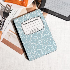 May include: A composition book with a light blue cover and a white abstract pattern. The cover has a white label with the text "COMPOSITION BOOK". The book measures 24.7 x 19.0 cm and contains 100 sheets. A Kindle e-reader is in the background.