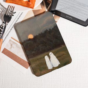 May include: A Kindle case featuring a landscape painting with a sunset, trees, and two white ghost figures. The case has a matte finish and is rectangular. The text "The cal" and "FERENCE BETWEEN YOU AND ME IS THAT YOU ARE WAITING F" is visible.
