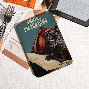 Pode incluir: Uma capa de livro turquesa com o texto "SHHH... I'M READING" apresenta um gato preto de óculos, lendo um livro. O gato está descansando em uma almofada listrada. Um leitor Kindle é visível no fundo.