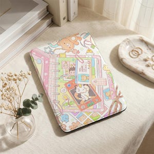 May include: A colorful, patterned tablet case featuring a cartoon design with a bear, stars, and other playful elements. The case has a pink, yellow, and white color scheme, with a spiral notebook design. A card with a cat illustration is visible.