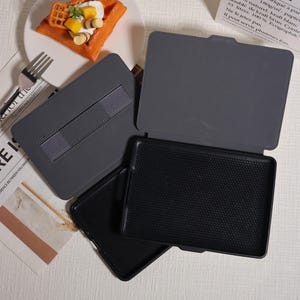 May include: A gray tablet case with a black interior and a textured surface. The case is open, revealing the interior. The case has a built-in stand. The background includes a plate with a waffle and a fork.