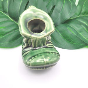 May include: A small, green ceramic boot-shaped planter with a glossy finish. The boot has a round opening at the top and is decorated with black stripes and dots. The planter is set against a backdrop of large, green tropical leaves.
