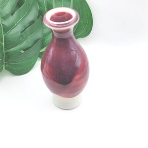 May include: A small, decorative vase with a deep burgundy glaze. The vase has a rounded body and a narrow neck with a white base. The rim of the vase is a lighter shade of burgundy. The vase is approximately 5 inches tall.