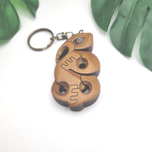 May include: Wooden tiki-style keychain with a bronze-colored key ring. The keychain is carved with a detailed design, featuring a face and abstract shapes. The wood has a natural, light brown color with darker accents. A small, iridescent inlay is visible.