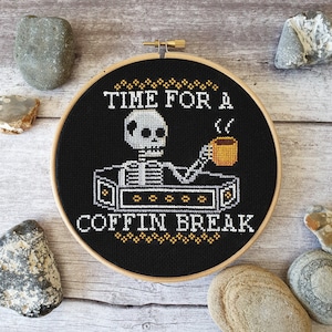 May include: A black and white cross-stitch embroidery hoop featuring a skeleton holding a cup of coffee. The text reads "Time for a Coffin Break".