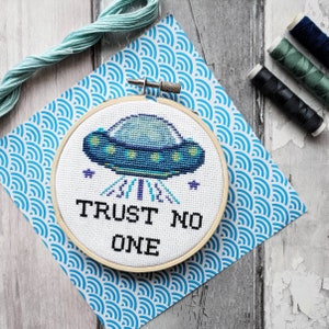 Könnte beinhalten: Ein weißer Stickrahmen mit einem blauen und grünen UFO und dem Text "TRUST NO ONE" auf einem blau-weiß gemusterten Hintergrund.