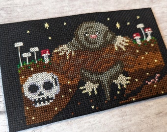 May include: Cross-stitch embroidery depicting a brown mole with a white belly, holding a skull in its paws. The mole is in a dark brown dirt mound with white mushrooms and a starry night sky.