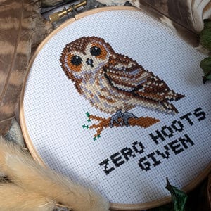 May include: Cross-stitch embroidery of an owl with brown, tan, and gray hues, set in a wooden hoop. The text "ZERO HOOTS GIVEN" is stitched below the owl. Feathers and wood pieces surround the hoop.