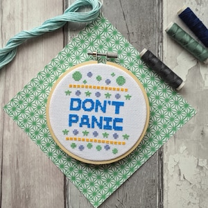 May include: A white embroidery hoop with a green, blue, and yellow cross-stitch design that reads "DON'T PANIC". The hoop is on a green and white patterned fabric.