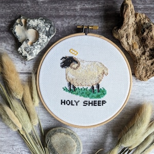 May include: A white sheep with a halo above its head is embroidered on a white canvas in a wooden embroidery hoop. The sheep is standing on green grass and the text "HOLY SHEEP" is written below the sheep.