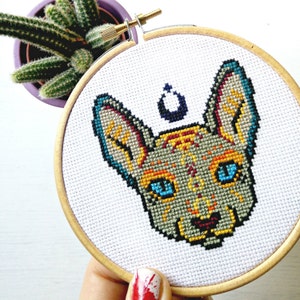 May include: A cross-stitch embroidery hoop with a colorful image of a Sphynx cat with blue eyes. The cat is surrounded by a white background with a small crescent moon above its head.