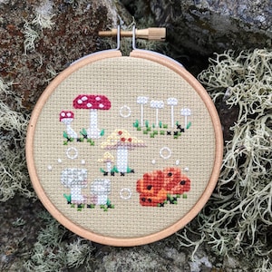May include: A beige embroidery hoop with a cross-stitch design of mushrooms. The design features red, white, and orange mushrooms with green leaves and white dots.
