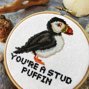 May include: A white embroidery hoop with a black and white cross-stitch design of a puffin. The text "You're a stud puffin" is stitched below the puffin.