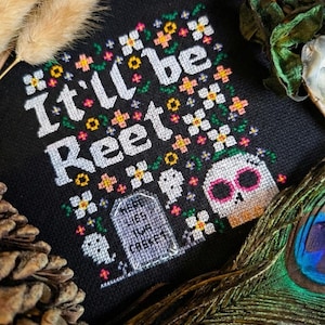 Peut inclure: Broderie au point de croix noire avec l'inscription blanche "It'll be Reet", ornée de motifs floraux et de têtes de mort. Une pierre tombale avec l'inscription "Here lies your casket" est également présente. Le tout est encadré de plantes séchées et d'une plume de paon.